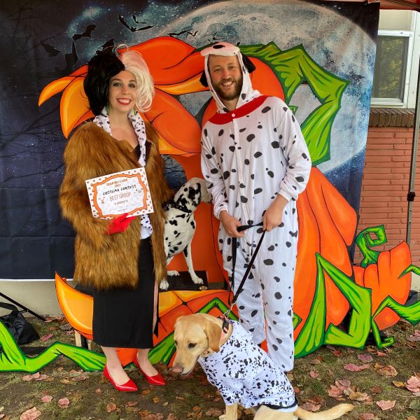 A woman dressed as Cruella De Ville poses with a man dressed as a dalmatian, and a yellow lab dressed as a dalmatian. Also an actual dalmatian.