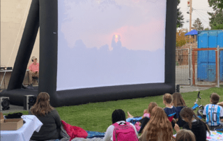 A large outdoor movie screen displays a romantic sunset scene as neighbors look on from park blankets and camp chairs.