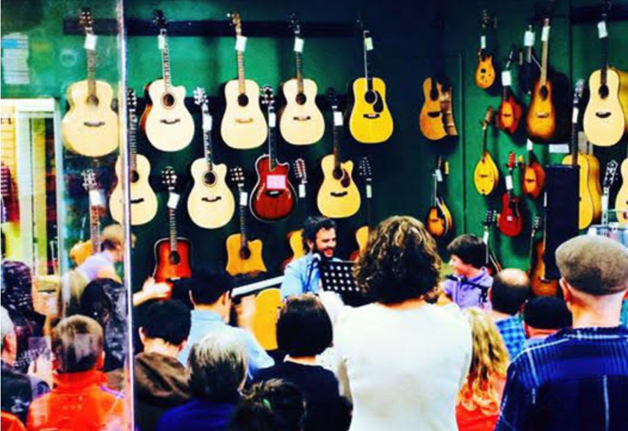 Vintage photo of Jared seated before a green wall lined with guitars surrounded by students as he kicks off a recital at the Guitar Store.